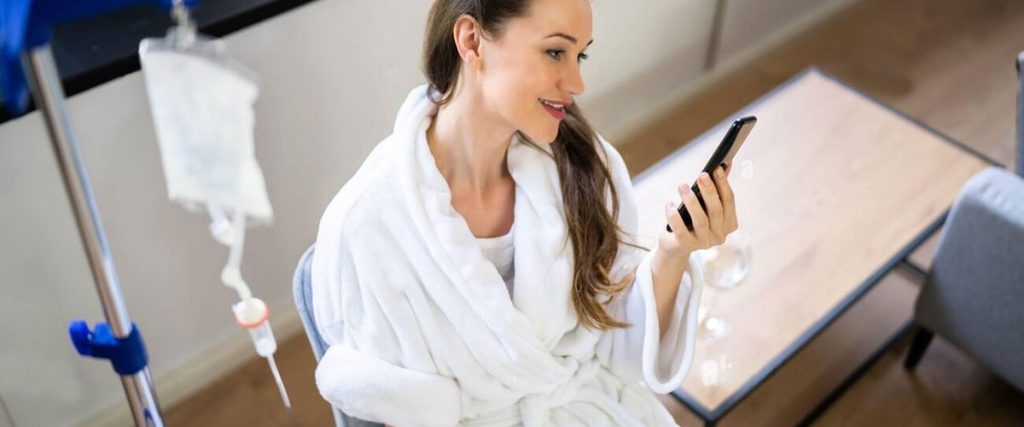 A woman in a white robe holding a cell phone while having an IV Drip Therapy from Sentral IVs - Your Partner in Hydration and Wellness.