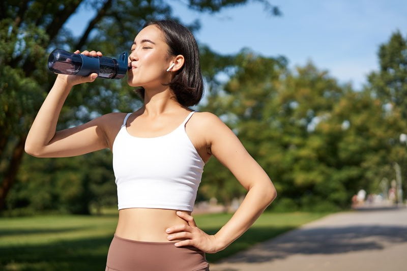 Smiling asian fitness girl drinks water, workout in park in Bali, stay hydrated during jogging training